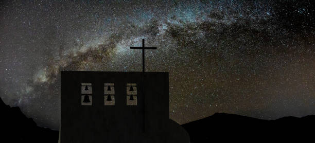 Cross on church over clear night sky and bright Milky Way . Six bells of different sizes on the top. Peaks of high mountains in the background. Night shot long exposure.