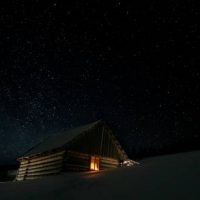 Stars in the night sky and a wooden house with a glowing window in winter