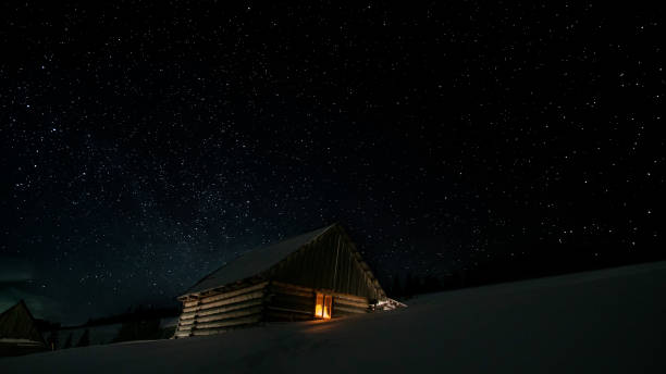 Stars in the night sky and a wooden house with a glowing window in winter